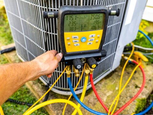 A person’s hand holds a digital manifold gauge attached to an outdoor HVAC unit with yellow, blue, and red hoses for AC troubleshooting. The gauge displays pressure and temperature readings to help diagnose AC common causes or an AC not cooling issue. | High 5 Plumbing, Heating & Cooling