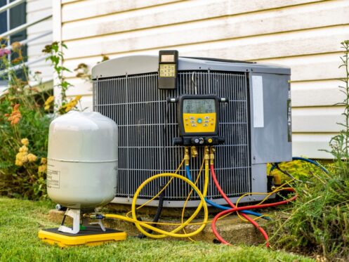 An air conditioning unit sits outside a house, connected to various hoses and diagnostic gauges. A refrigerant tank is nearby on the grass. The unit is next to a white siding wall with some plants visible around it. | High 5 Plumbing, Heating & Cooling