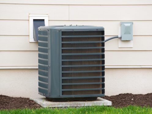A central air conditioning unit sits on a concrete pad outside a house, with electrical wiring connected to a nearby box on the exterior wall. AC units are known for using the most electricity, while the ground is covered with mulch and grass. | High 5 Plumbing, Heating & Cooling