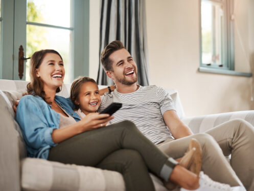 A woman, a man, and a young girl sit together on a couch in a living room, smiling and looking at something off-camera. The woman holds a remote control in their smart home, filled with natural light from energy-efficient windows behind them. | High 5 Plumbing, Heating & Cooling