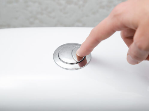 A close-up of a hand pressing a chrome dual flush button on the top of a white toilet tank. | High 5 Plumbing, Heating & Cooling
