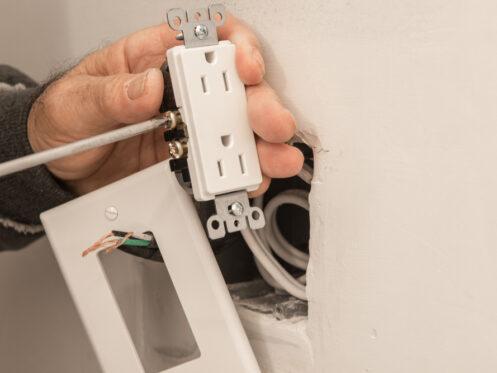 A person is converting a wall fixture by installing a white outlet using a screwdriver. The outlet cover and wires are visible, and the unfinished wall shows where the light switch or outlet is being fitted. | High 5 Plumbing, Heating & Cooling