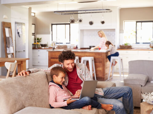 A man and a boy sit on a couch looking at a laptop, both smiling—perhaps researching installing a backup generator for winter. In the background, a woman and girl are at the kitchen island in their bright, modern open-plan living space. | High 5 Plumbing, Heating & Cooling