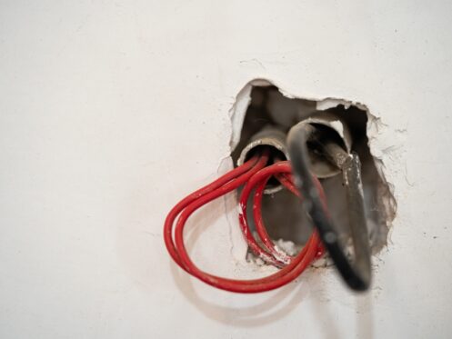 A close-up of an electrical wall box shows exposed electrical wires—red and black—protruding from a rough hole in a white plaster wall, highlighting the need for electrical safety as the wires await connection to fixtures or covers. | High 5 Plumbing, Heating & Cooling