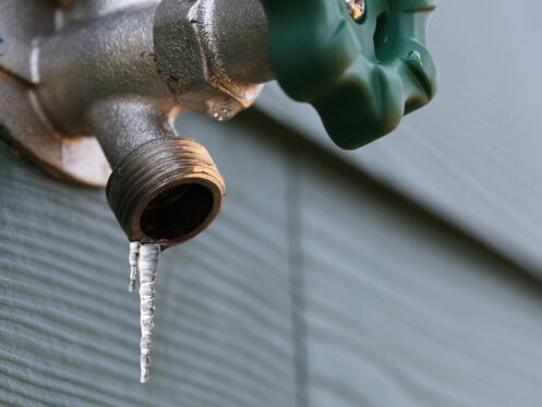 A close-up of an outdoor metal water faucet with a green handle, attached to a wall, with a small icicle hanging from the spout—an early sign of winter and a reminder of the risk of frozen pipes. | High 5 Plumbing, Heating & Cooling