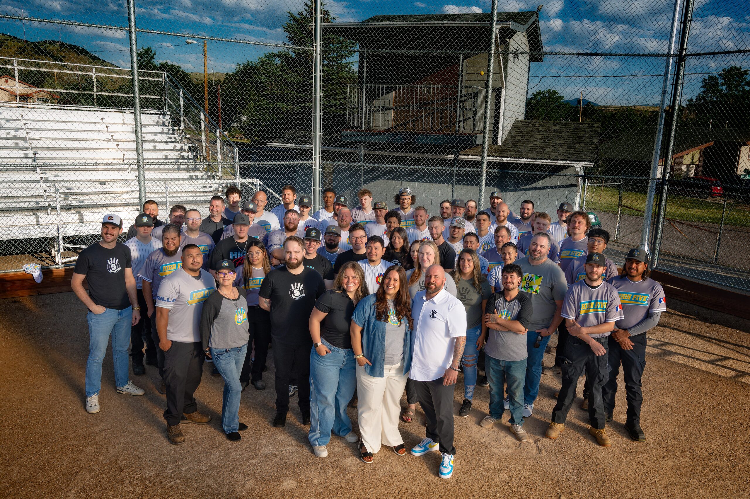A group of about 50 people pose together on a baseball field in front of bleachers and a chain-link fence. Most are wearing t-shirts and jeans. It is sunny, and the group stands in a semicircle, smiling at the camera. | High 5 Plumbing, Heating & Cooling