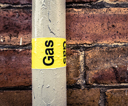 A gray pipe with a yellow "Gas" label runs vertically in front of a brick wall, showing water droplets on its surface—a sign that gas line repair may be necessary. | High 5 Plumbing, Heating & Cooling
