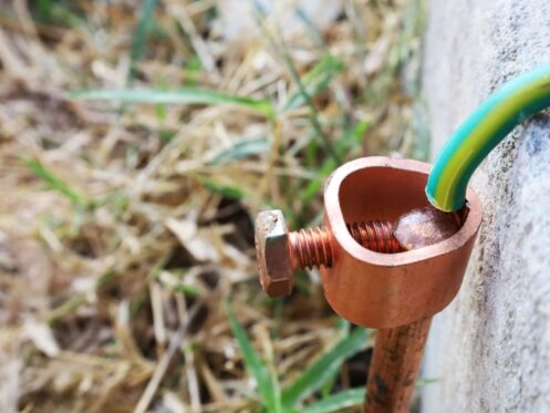 A close-up view of a copper grounding rod with a green and yellow ground wire attached by a metal clamp, positioned next to a concrete surface and surrounded by grass, highlights the importance of electrical grounding for safety. | High 5 Plumbing, Heating & Cooling