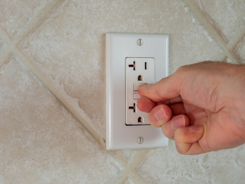 A hand is pressing the test button on a white GFCI outlet mounted on a tiled wall, demonstrating basic GFCI troubleshooting when an outlet keeps tripping. | High 5 Plumbing, Heating & Cooling