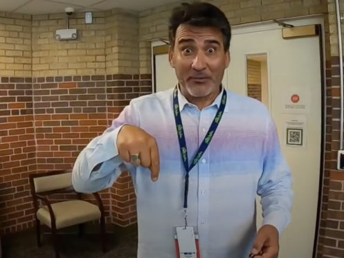 A man wearing a light-colored button-up shirt and lanyard stands indoors near brick walls, pointing downward with a surprised facial expression. A chair and an open door are visible in the background. | High 5 Plumbing, Heating & Cooling