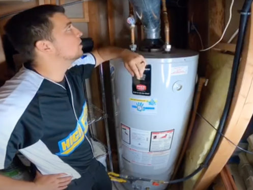 A man in a black and white shirt stands next to a water heater in a utility room, resting his arm on top of the unit and looking up at the pipes and connections above it. | High 5 Plumbing, Heating & Cooling