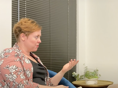 A person with short, light brown hair and a floral patterned cardigan gestures with one hand while seated indoors near a small table with a potted plant. Black blinds cover the window behind them. | High 5 Plumbing, Heating & Cooling