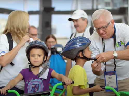 An adult helps a young boy fasten his bicycle helmet while a girl wearing a helmet stands nearby; other adults and children are present in the background. | High 5 Plumbing, Heating & Cooling