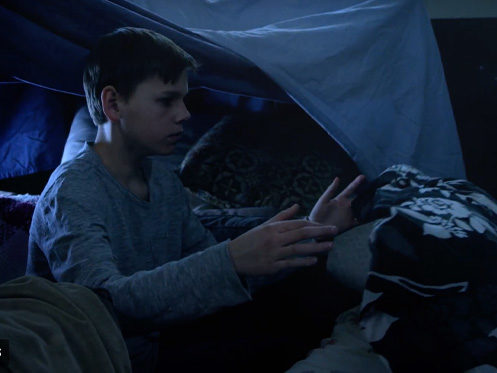 A boy wearing a gray long-sleeve shirt sits inside a dimly lit blanket fort, reaching forward with both hands toward a pile of pillows and blankets. | High 5 Plumbing, Heating & Cooling