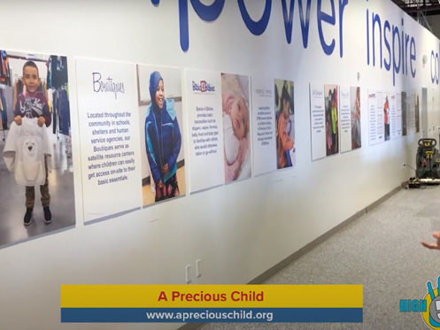 A wall display features large photos and informational posters about community services. The wall has words like “power” and “inspire.” The bottom of the image shows a “A Precious Child” banner with a website link. | High 5 Plumbing, Heating & Cooling
