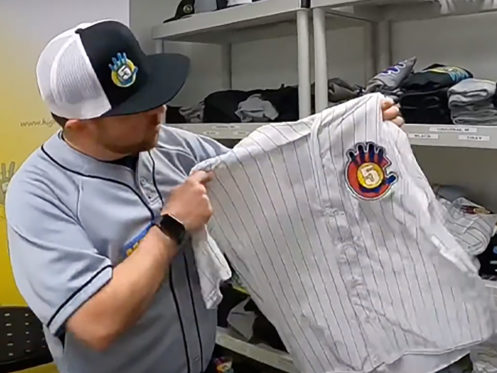 A man wearing a baseball jersey and cap holds up a white pinstriped baseball shirt with a colorful "5" logo patch, standing in front of shelves with folded clothes. | High 5 Plumbing, Heating & Cooling