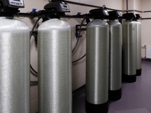Five large industrial water filtration tanks with digital control panels are lined up against a wall, interconnected by pipes and hoses in a clean, tiled utility room. | High 5 Plumbing, Heating & Cooling