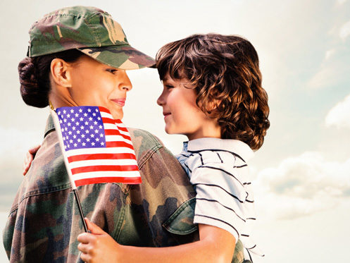 A woman in military uniform holds a young boy in her arms. The boy holds a small American flag, and they look at each other, both smiling. The background is mostly sky with clouds. | High 5 Plumbing, Heating & Cooling