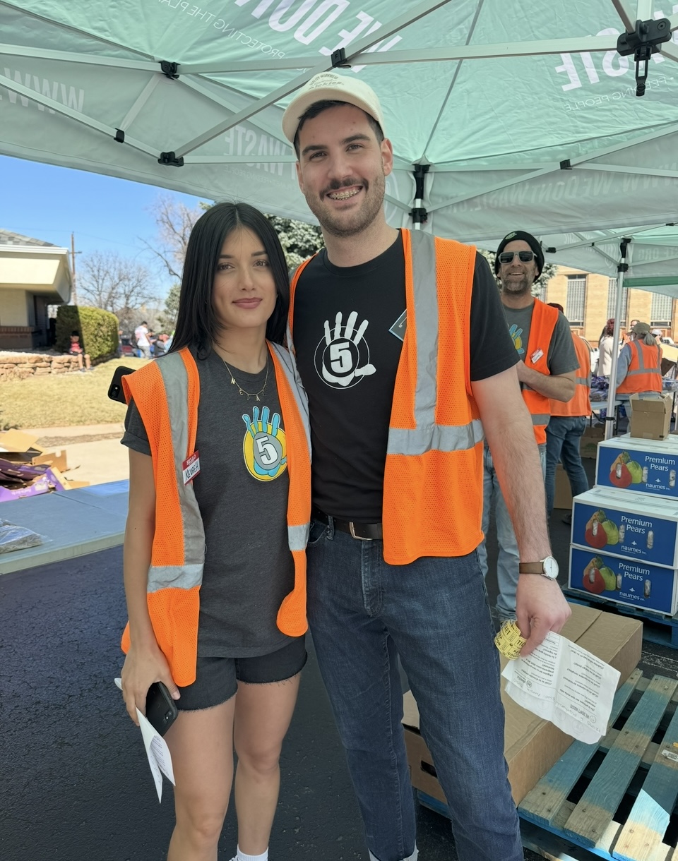 A group of people wearing masks stand behind large blue and yellow bins, holding up plastic-wrapped items at a High 5 Cares event in a warehouse filled with shelves of boxes in the background. | High 5 Plumbing, Heating & Cooling
