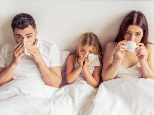 A man, a young girl, and a woman sit together in bed, holding tissues to their noses as if sneezing, likely due to dry winter air in Denver. All three are under white blankets and wearing white tops. | High 5 Plumbing, Heating & Cooling