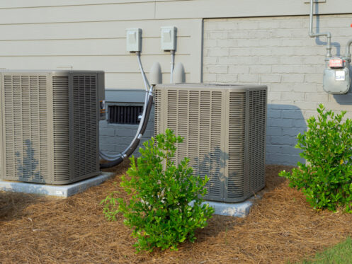 Two outdoor air conditioning units sit on concrete pads beside a beige house with a brick foundation. Small green shrubs and mulch provide neat AC unit landscaping, creating an attractive, low-maintenance look for the area. | High 5 Plumbing, Heating & Cooling
