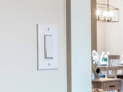 A white dimmer switch on a beige wall in the foreground; in the background, a dining area with a table, chairs, and a chandelier is visible. | High 5 Plumbing, Heating & Cooling