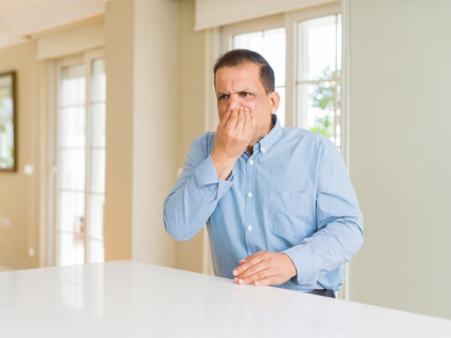 A man in a blue button-up shirt sits at a white table indoors, holding his nose with a displeased expression, as if reacting to a sewer gas smell. Large windows and light-colored walls are visible in the background. | High 5 Plumbing, Heating & Cooling