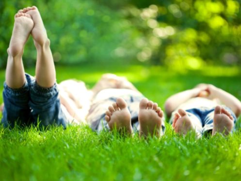 Three children lie barefoot on green grass, their feet in the foreground near an outdoor faucet. The blurred greenery and sunlight in the background evoke a relaxed outdoor setting. The children are wearing jeans. | High 5 Plumbing, Heating & Cooling