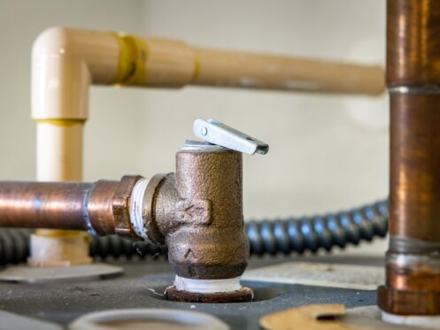 Close-up of a water heater pressure relief valve attached to a copper pipe, with additional plastic and metal pipes in the background. The valve is installed on a flat metal surface. | High 5 Plumbing, Heating & Cooling