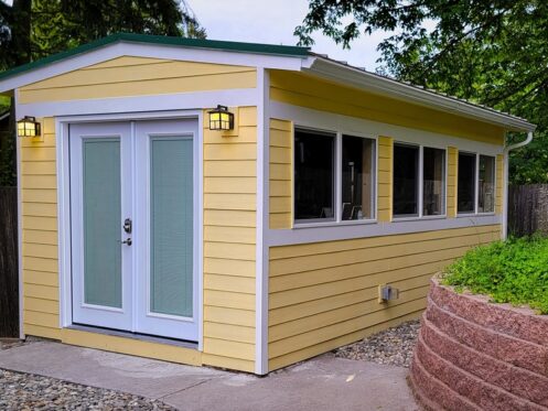 A small yellow power shed with white trim, double glass doors, and several windows sits on a concrete path, surrounded by a wooden fence and a landscaped area with a stone retaining wall and greenery. | High 5 Plumbing, Heating & Cooling