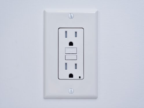 A white electrical outlet with two sockets and two rectangular buttons, mounted on a white wall. This tamper-resistant GFCI outlet offers enhanced safety features to help protect against electrical shocks. | High 5 Plumbing, Heating & Cooling