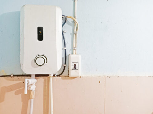A white wall-mounted tankless water heater with visible pipes and a control panel is installed on a pale blue and beige tiled wall. An electrical switch is positioned next to the heater, highlighting easy access for maintenance. | High 5 Plumbing, Heating & Cooling