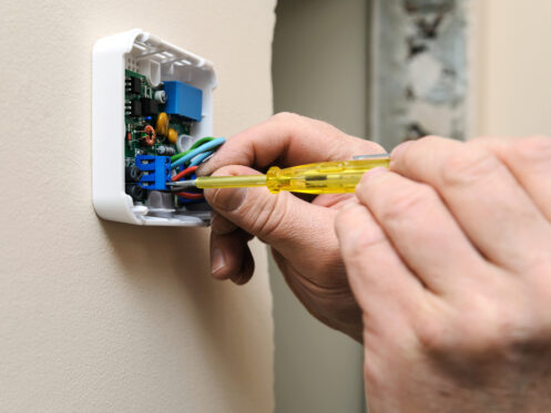 A person uses a screwdriver to adjust wires inside a wall-mounted electronic device or switch box, referencing thermostat wire color codes, with exposed circuits and components visible. | High 5 Plumbing, Heating & Cooling