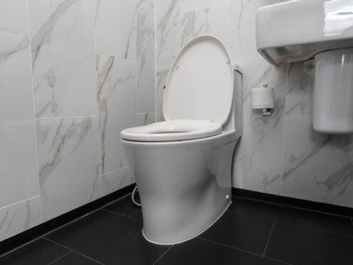 A modern white toilet with the lid open—ideal for toilet troubleshooting like when the toilet tank is not filling—sits next to a wall-mounted sink and toilet paper holder in a bathroom with marble-patterned walls and a black tiled floor. | High 5 Plumbing, Heating & Cooling