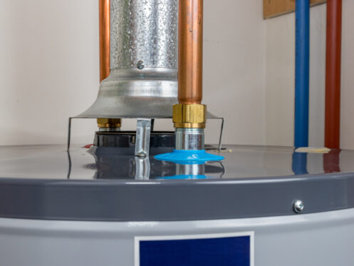 A close-up view of the top of a gas water heater shows metal and copper pipes, a vent, and a blue plastic cap, with a white wall in the background. | High 5 Plumbing, Heating & Cooling