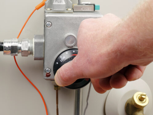 A hand turns the temperature control dial on a water heater valve, highlighting water heater energy usage, with "Pilot" labeled above and pipes attached to the unit. | High 5 Plumbing, Heating & Cooling