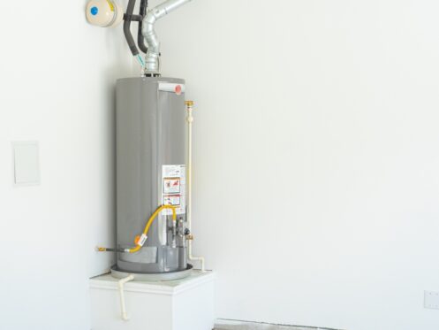 A gray water heater is installed in the corner of a white room, elevated on a small platform, with pipes and ventilation attached. The unfinished room highlights the appliance, raising questions about water heater longevity in such spaces. | High 5 Plumbing, Heating & Cooling