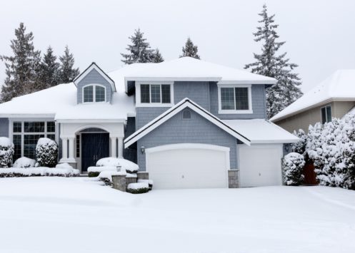 A two-story suburban house with snow covering the roof, driveway, and yard. Evergreen trees and bushes are blanketed in snow, while the overcast sky hints at a recent snowfall—perfect weather to check and winterize plumbing systems. | High 5 Plumbing, Heating & Cooling