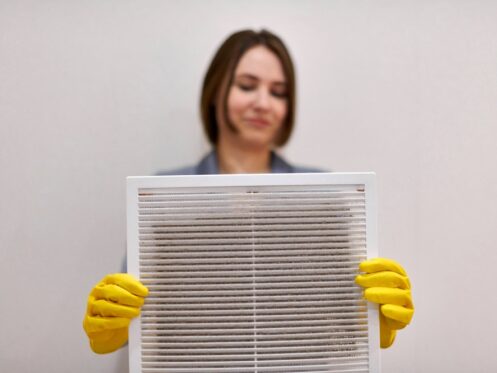 A person wearing yellow cleaning gloves holds up a dirty air filter, showing dust and debris that impact air filter efficiency. The background is plain and out of focus. | High 5 Plumbing, Heating & Cooling