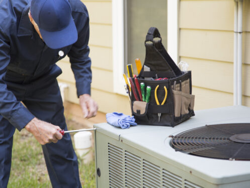 A person in a navy uniform and cap uses a screwdriver for air conditioner troubleshooting on an outdoor unit. A tool bag with various tools and a blue cloth sits on top of the unit next to them. | High 5 Plumbing, Heating & Cooling