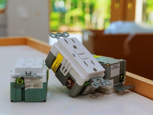 Three electrical outlets, including a GFCI outlet for Load vs Line Wiring difference, rest on a white surface in a room under construction with blurred green scenery visible through windows in the background. | High 5 Plumbing, Heating & Cooling