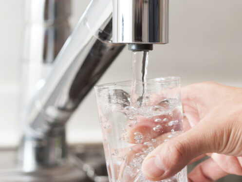 A hand holds a clear glass under a modern kitchen faucet, filling it with water filtered by a whole-home water filtration system. The background shows part of a stainless steel sink and countertop. | High 5 Plumbing, Heating & Cooling