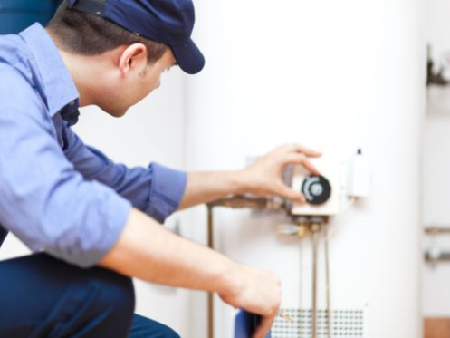 A person wearing a blue uniform and cap is performing maintenance by adjusting the control panel on a water heater in an indoor basement setting. | High 5 Plumbing, Heating & Cooling