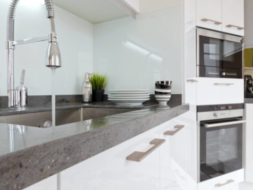 Modern kitchen with a gray countertop, stainless steel sink, and chrome pull-down faucet. Stacked white plates and bowls sit by small plants—perfect for those looking to improve water quality with stylish fixtures in white cabinetry. | High 5 Plumbing, Heating & Cooling