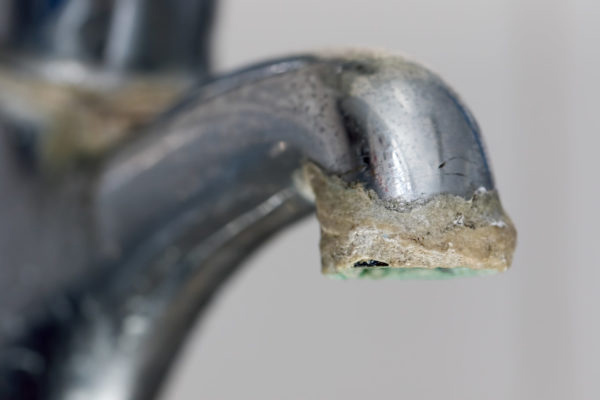 Close-up of the end of a metal faucet covered in thick, crusty white and yellow limescale buildup. The tarnished faucet with heavy mineral deposits highlights the need for professional Piping & Repiping Services. | High 5 Plumbing, Heating & Cooling