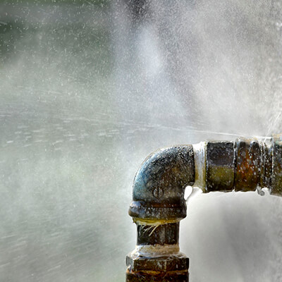 A close-up of a metal pipe joint with water spraying out from a leak, creating a mist around the area. The background is blurred and out of focus, highlighting the need for expert Piping & Repiping Services. | High 5 Plumbing, Heating & Cooling