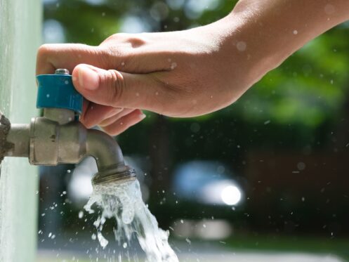 A person’s hand is turning on a blue-handled outdoor water faucet, with water flowing out of the tap and droplets splashing—highlighting reliable access, much like gas plumbing during a power outage. The background is blurred greenery and light. | High 5 Plumbing, Heating & Cooling
