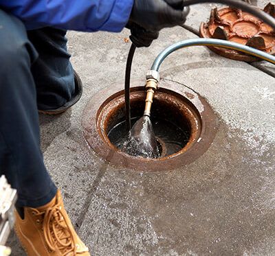 A person wearing a blue jacket, black gloves, and tan work boots is performing sewer repair, holding a hose and inserting it into an open drain or manhole on a concrete surface. Water and equipment are visible around the opening. | High 5 Plumbing, Heating & Cooling