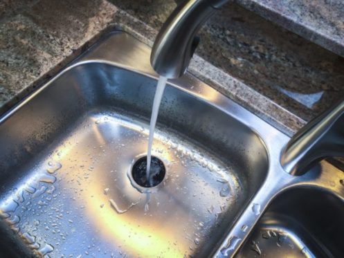 A kitchen sink with water running from a faucet directly into the drain, reminding you to practice plumbing protection—especially after rinsing Thanksgiving foods. Some water droplets are visible on the stainless steel surface around the drain. | High 5 Plumbing, Heating & Cooling