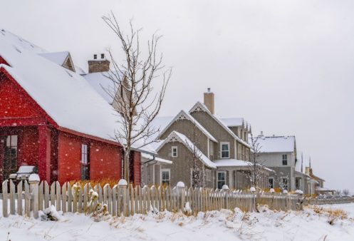 Several houses, including a prominent red one, are lined up behind a white picket fence on a snowy day. Snow blankets the roofs and ground—an ideal reminder to take steps for pipe freezing prevention this winter. | High 5 Plumbing, Heating & Cooling